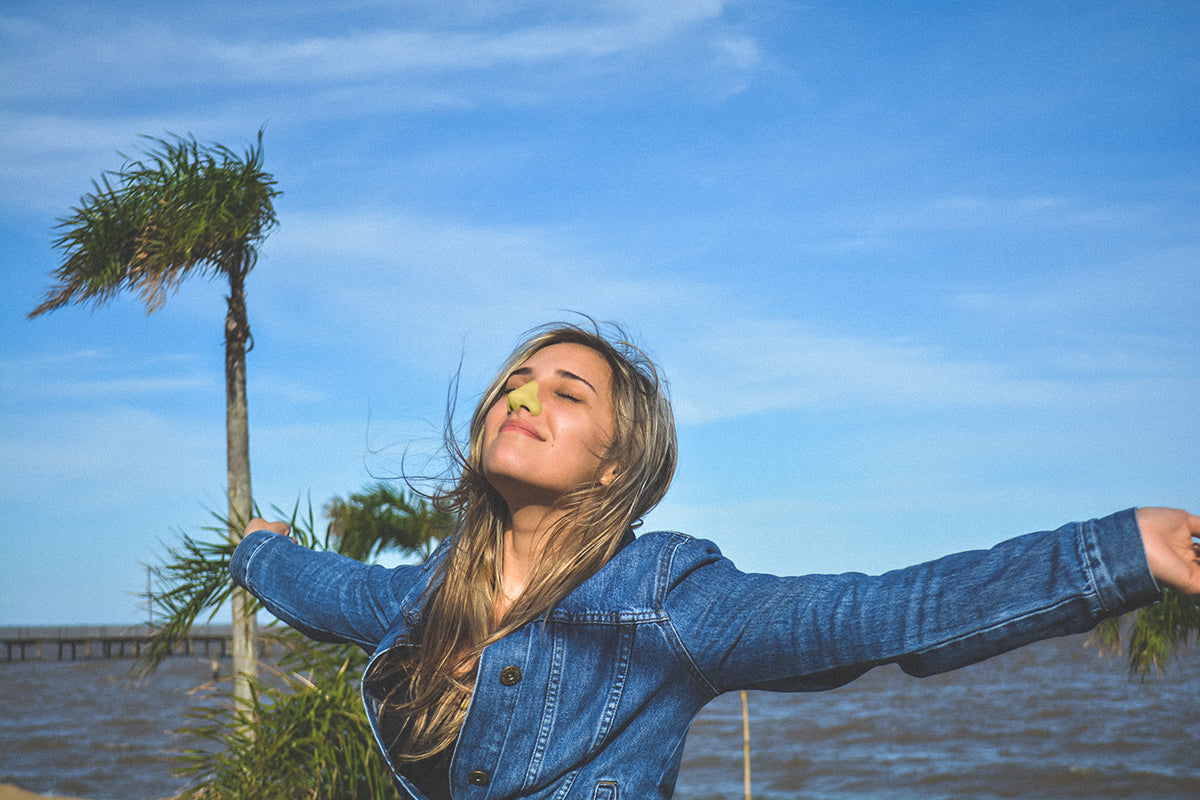 Woman standing with eyes closed and arms spread out with Nöz's yellow sunscreen on nose. 